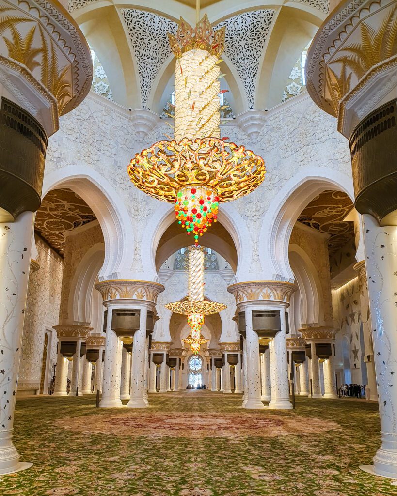 Inside the mosque in Abu Dhabi, the UAE which has rounded white and yellow domed roofs and columns. Hanging from the roof are three huge chandeliers made of white, red, orange and green jewels and a big gold plate going around all three of them. Underneath them is a huge green, red and yellow carpet that takes up the entire floor "Nine of the Best Budget Friendly Ways to see Dubai"