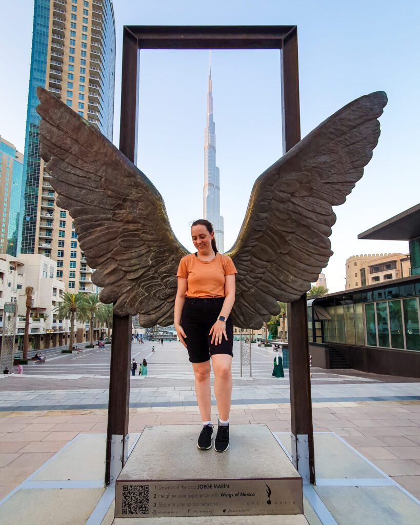 Krissie standing in front of a sculpture of a giant pair of bronze wings with a big rectangular frame behind them that perfectly frame the Burj Khalifa near the Dubai Mall in Dubai, UAE. Around the sculpture are some more buildings and palm trees "Nine of the Best Budget Friendly Ways to see Dubai"