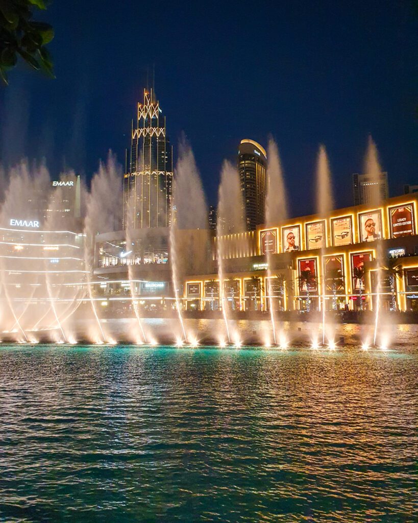 Part of the outside of Dubai Mall in Dubai, UAE, lit up at night. In front of it is a fountain with many water spouts shooting up into the sky, also lit up. In front of those is a body of water and the sky is dark and clear "Nine of the Best Budget Friendly Ways to see Dubai"