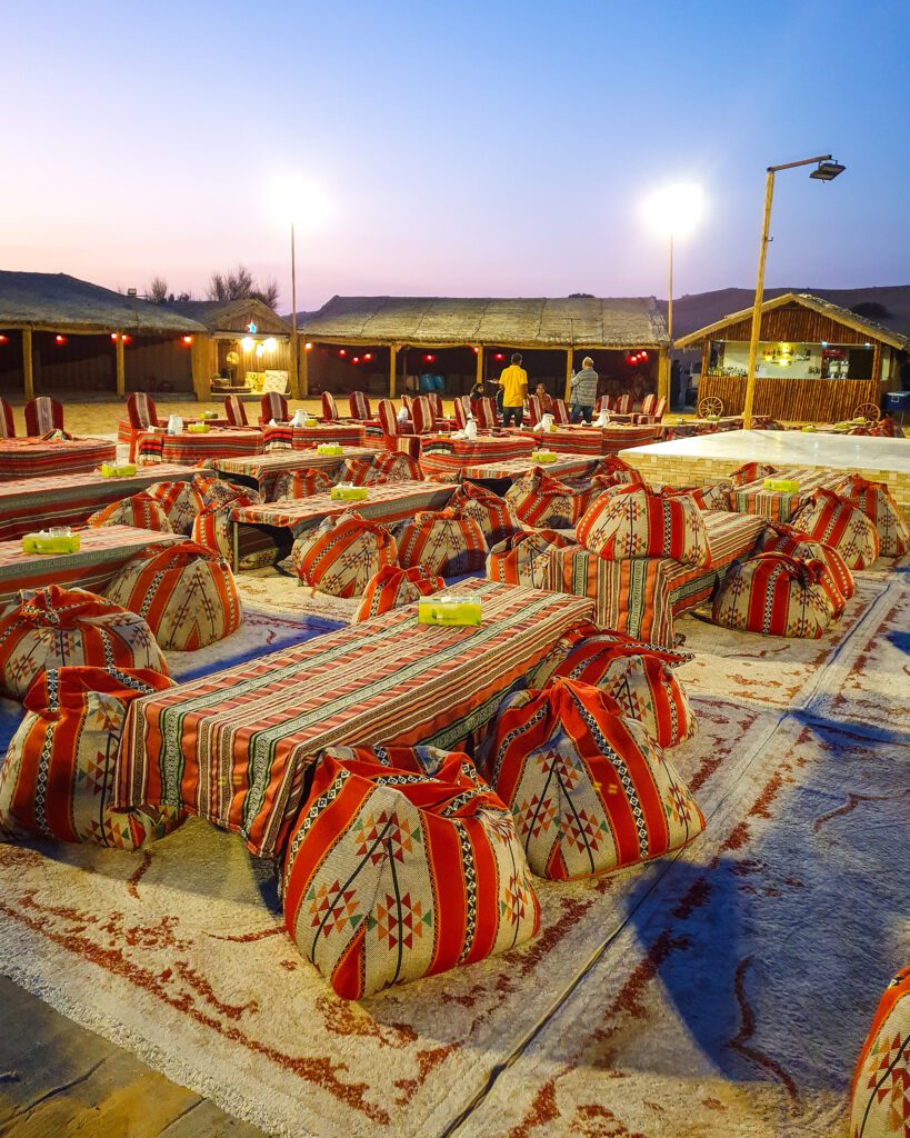 An outdoor seating area with tables covered in patterned table cloths and lots of patterned bean bags around the tables. Under them are rubs and around the tables are undercover spaces and some lampshades flooding light onto the area as the sun sets in the Arabian Desert in the UAE "Nine of the Best Budget Friendly Ways to see Dubai"