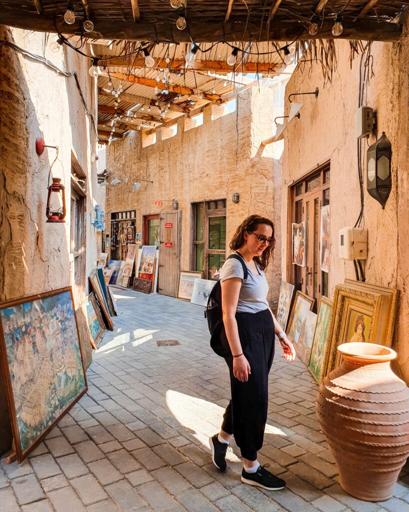 Krissie standing looking at a big terracotta pot in the historical neighbourhood in Dubai, UAE. Behind her is a narrow sandstone coloured street with walls all the way up to a covering over it all. Along the floor leaning on the walls on both sides are paintings and above are some string lights "Nine of the Best Budget Friendly Ways to see Dubai"