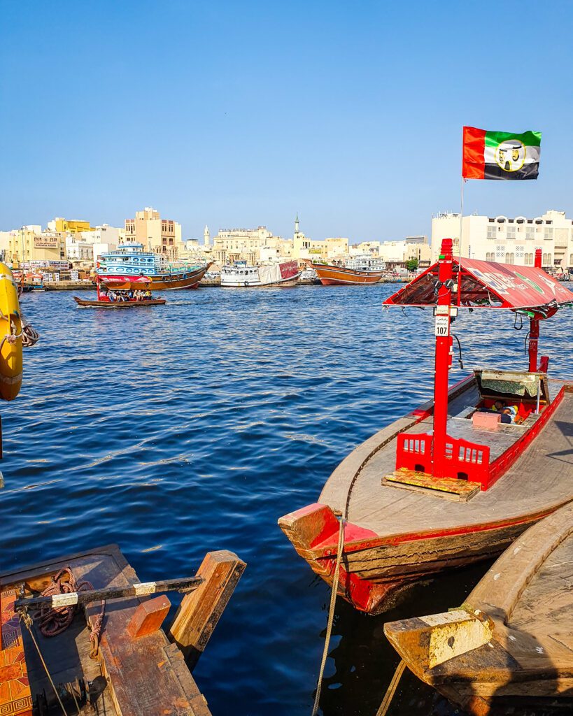 The beautiful blue water of Dubai Creek in the UAE with small wooden boats, one of them with the UAE flag on it. Behind the boat in the foreground are some white and cream buildings and more wooden boats under a clear blue sky "Nine of the Best Budget Friendly Ways to see Dubai"