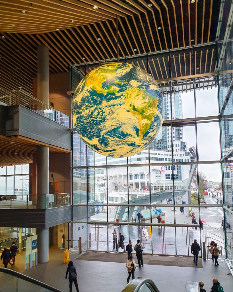 Inside a building in Vancouver, Canada. The building has a big glass window with doors down the bottom, and on the left you can see 2 levels of the building. Hanging from the roof in the middle of the photo is a giant model of the Earth lit up yellow and there's people walking around under it "Plan Your Visit: The Best Christmas Markets In Canada"