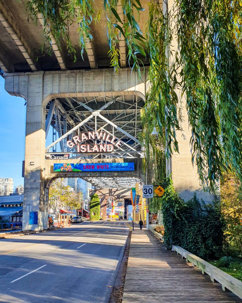 A road under a road bridge leading onto Granville Island in Vancouver, Canada. There's a big sign that says Granville Island above the road and on the right are some plants and a path also leading to the island "Plan Your Visit: The Best Christmas Markets In Canada"