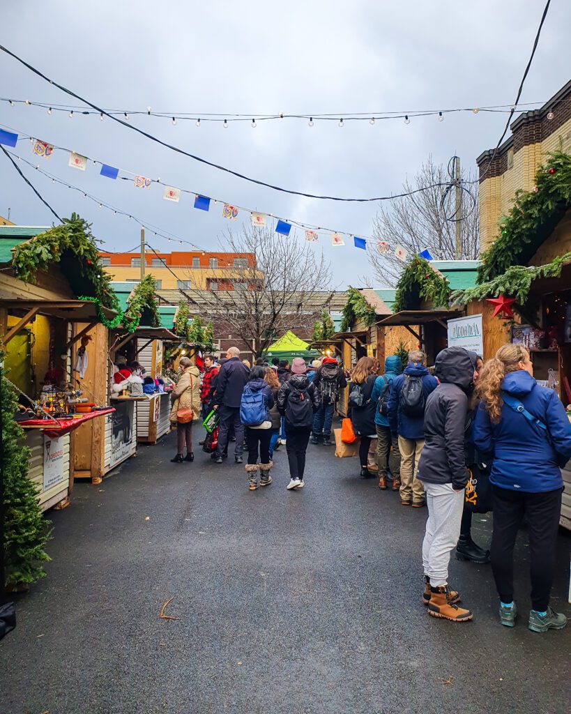 People walking through the Christmas village in Montreal, Canada. On either side of the path people are walking are wooden hugs with green plants on top and city buildings in the background "Plan Your Visit: The Best Christmas Markets In Canada"