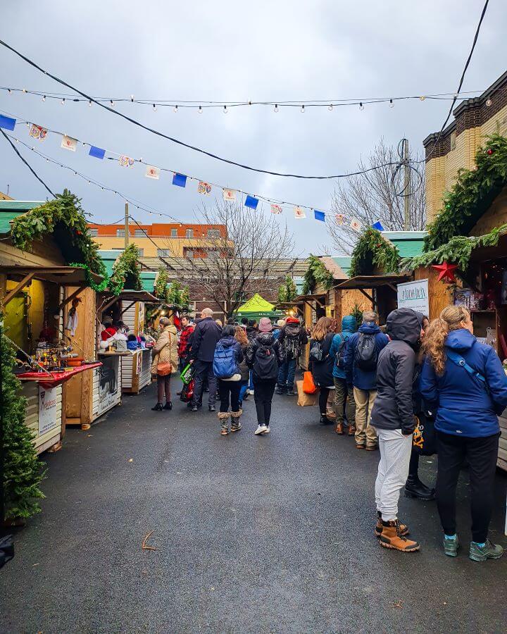 People walking through the Christmas village in Montreal, Canada. On either side of the path people are walking are wooden huts with green plants on top and city buildings in the background "Eight Great Free Things To See In Montréal"