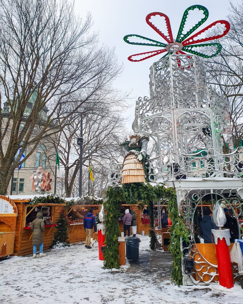 Inside part of the Christmas Market in Québec city, Canada. On the left are some wooden huts selling Christmas goodies with green plants and snow on top. Above the huts are some brown trees and a building outside the market and on the right is a big metal structure for people to stand under with green plants and big metal candle sculptures and on the top of it is a red and green flower looking structure. There's snow on the ground and people walking around the market "Plan Your Visit: The Best Christmas Markets In Canada"