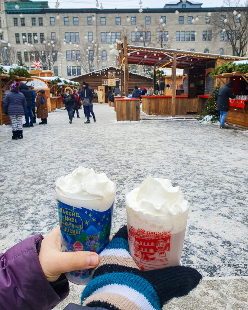 Krissie's hand and Katherine's gloved hand holding red and blue plastic cups with hot chocolates with whipped cream on top. Behind the cups is part of the Christmas Market in Québec city, Canada, with wooden German style huts, lights strung up, people walking around and snow on the ground. So magical! "Plan Your Visit: The Best Christmas Markets In Canada"