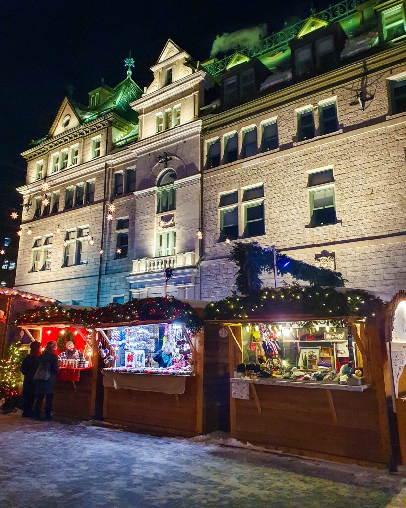 Plan Your Visit: The Best Christmas Markets In Canada. Three wooden German style huts with green plants and Christmas lights on top at the Québec city Christmas Markets in Canada. Inside the huts they're selling clothes and food and behind the huts is a huge stone building with a green roof. The photo is also taken at night so everything is lit up and there's a bit of snow on the ground.