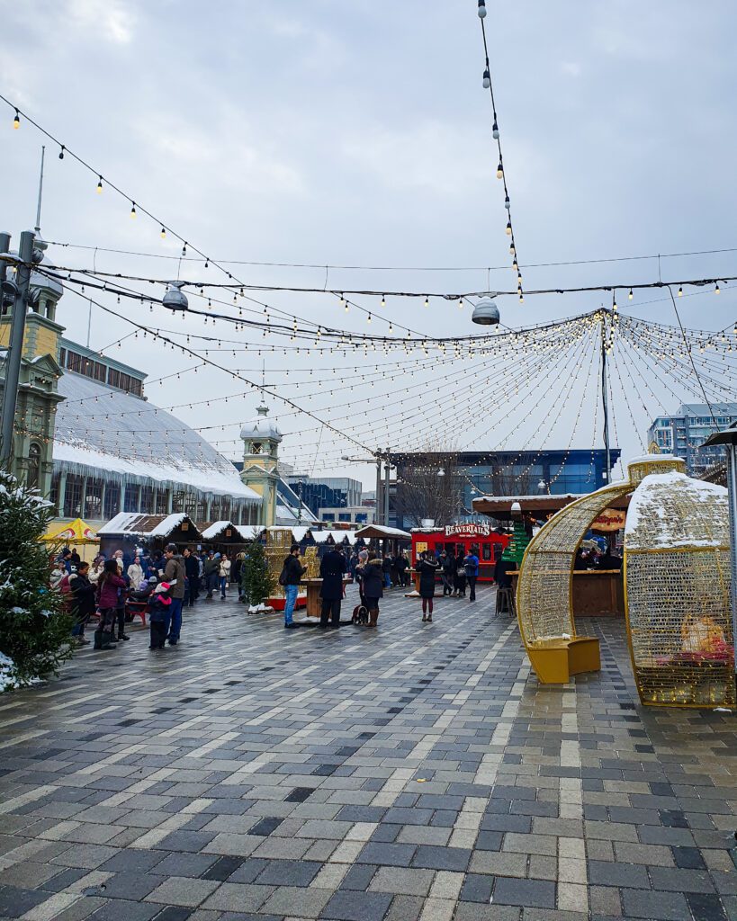 A big paved section at the Christmas Market in Ottawa, Canada. There are wooden huts around the edge of the paving with snow on top and on the right is a pole with many strings of lights coming off it above the huts. On the left is a little Christmas bauble style seating area and on the left is the Aberdeen Pavilion which is a big yellow building with a big grey roof "Plan Your Visit: The Best Christmas Markets In Canada"