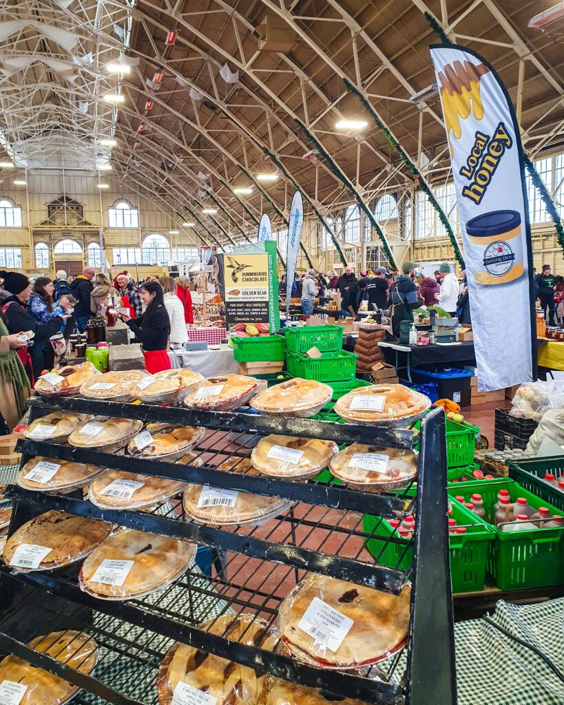 Inside the Aberdeen Pavilion in Ottawa, Canada. It has a high arched roof and is packed with people inside with a shelf of big fresh pies and a sign saying "local honey" and a bunch of green crates holding local produce "Plan Your Visit: The Best Christmas Markets In Canada"