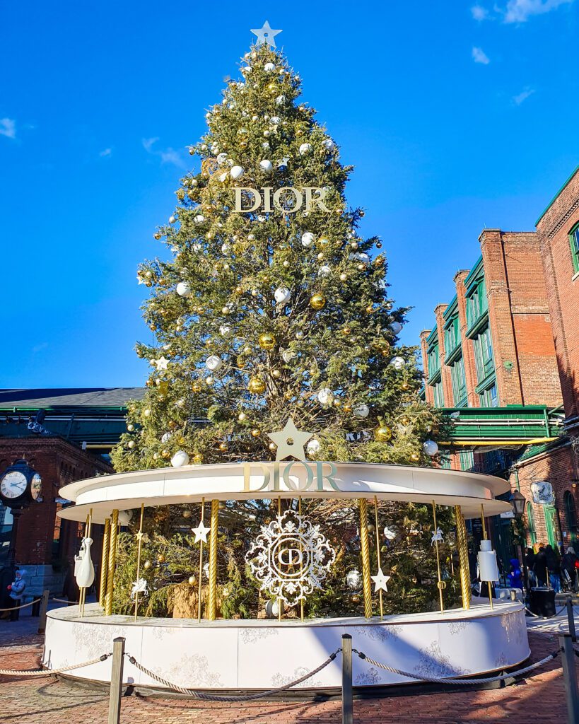 A big green Christmas tree at the Christmas Market in Toronto, Canada, with yellow and white baubles on it and a sign that says DIOR with a white platform at the bottom that also says DIOR. Around the tree is a rope barrier and behind it is a brick building with the blue sky above it "Plan Your Visit: The Best Christmas Markets In Canada"