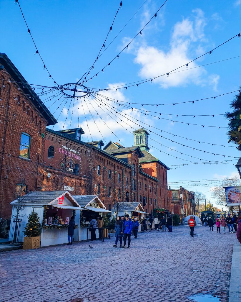 The distillery Christmas market in Toronto, Canada. The ground is paved and there's a big brick building on the left with a sign on it that says "Countdown to Christmas, 7 days". On the paved section are a couple of wooden huts selling Christmas goodies with people walking around and the blue sky above it all "Plan Your Visit: The Best Christmas Markets In Canada"