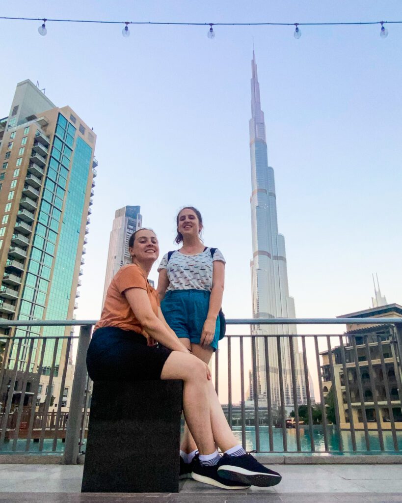 Nine of the Best Budget Friendly Ways to see Dubai. Krissie and Katherine posing in front of a railing, and behind it is the Burj Khalifa in Dubai, UAE, which has a thick base and turns into a small point way up in the sky. Around the Burj are other buildings and a shallow blue body of water.