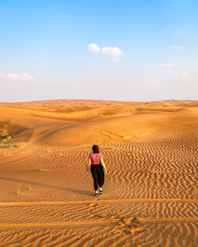 Krissie with a red shirt and black pants walking away from the camera in the Arabian Desert in the UAE. The entire photo is just hills of orange sand with some yellow grass and a blue sky above it all "Nine of the Best Budget Friendly Ways to see Dubai"