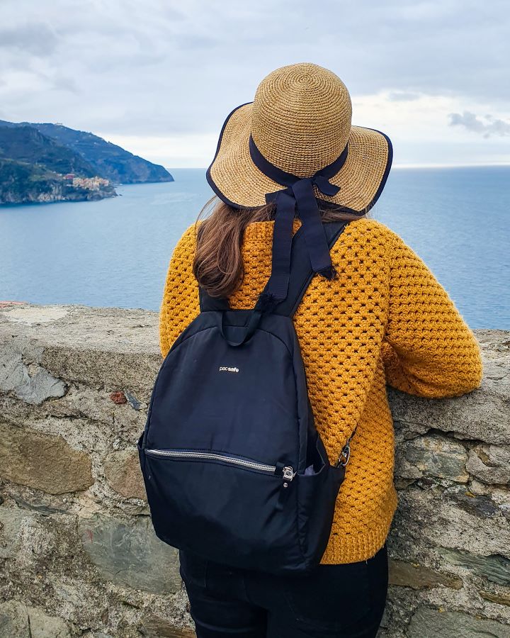 Krissie standing at a stone wall with a yellow jumper and straw hat on looking out at a view of the ocean and hills in Italy. On her back is her small Pacsafe bag with a zip on the front "The Ultimate Travel Packing Guide: 39 Essentials for Every Trip"