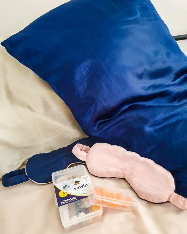 Blue and beige silk pillowcases on pillows with pink and blue silk eye masks in front of them. In front of those is a clear plastic case with some foam orange ear plugs in it and next to it is a small clear plastic sewing kit "The Ultimate Travel Packing Guide: 39 Essentials for Every Trip"