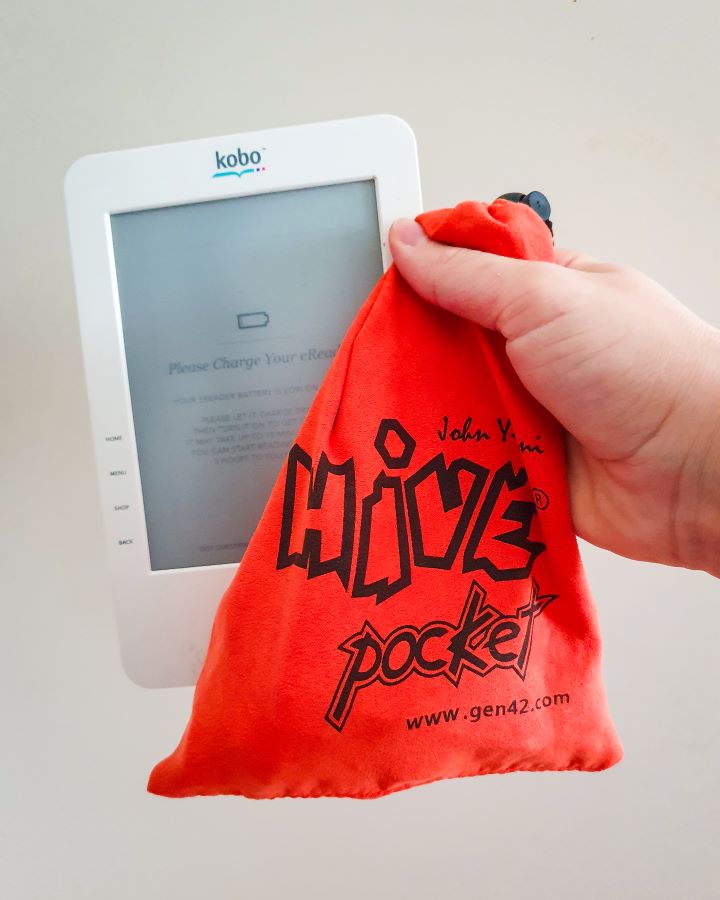 A hand holding a small white Kobo e-reader that says "Please charge your e-reader" on the screen and an orange bag that says "Hive pocket" on it "The Ultimate Travel Packing Guide: 39 Essentials for Every Trip"