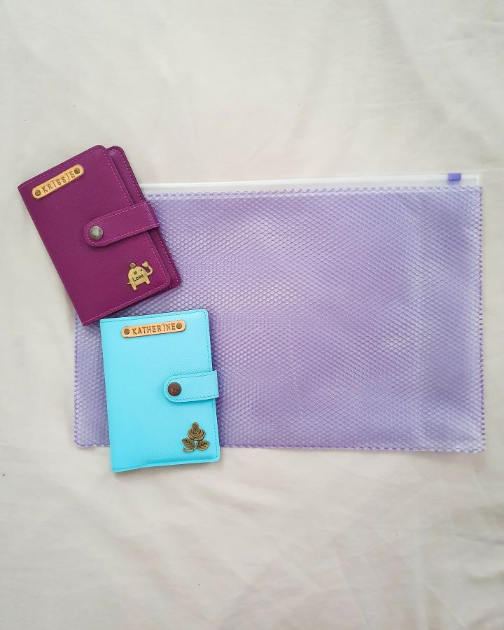 A clear plastic purple document holder with two passport holders on it. One is purple and says Krissie with a little elephant charm on it and one is blue and says Katherine with a little rose charm on it "The Ultimate Travel Packing Guide: 39 Essentials for Every Trip"