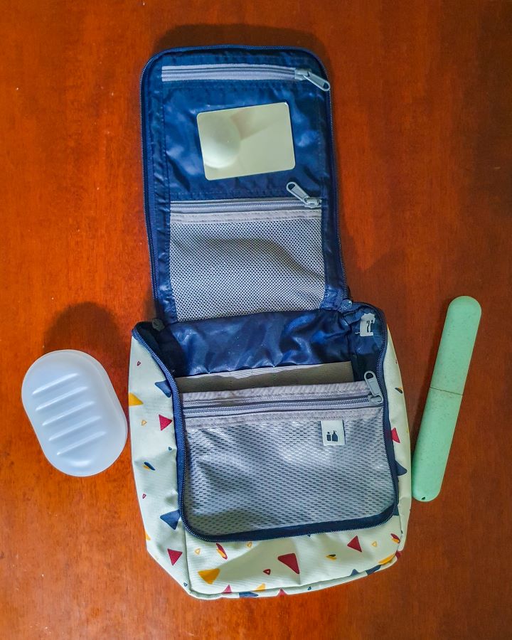 A waterproof toiletries bag lying open on a table. Inside are some mesh zip pockets and a little mirror. Next to it is a clear plastic soap holder and a green plastic toothbrush holder "The Ultimate Travel Packing Guide: 39 Essentials for Every Trip"