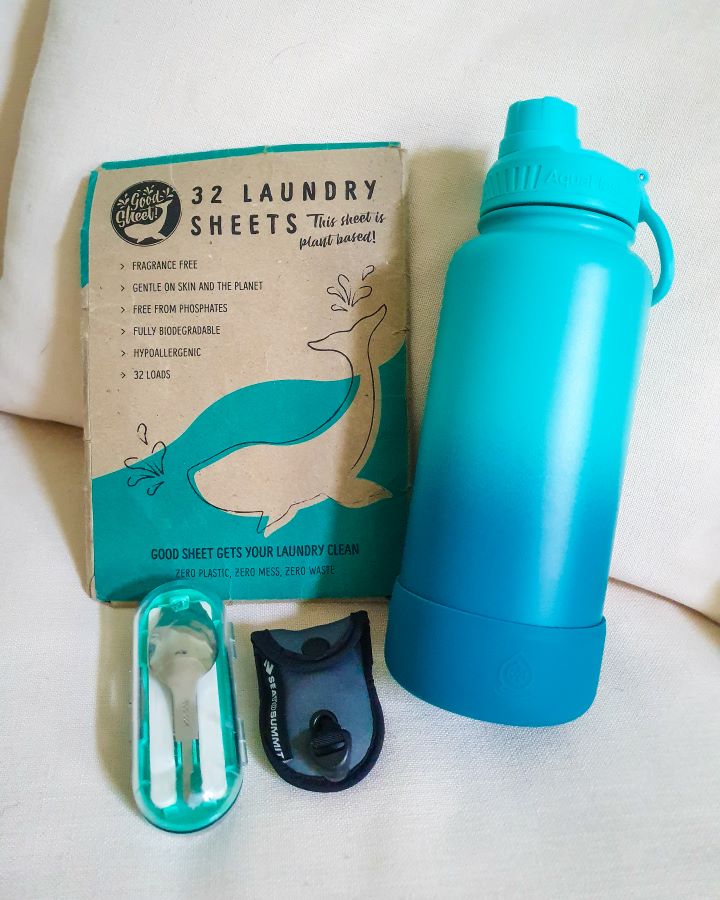 A big blue insulated water bottle with a packet of 32 laundry sheets next to it that looks a bit battered. Below them is a clear travel cutlery case with a little spoon head in it, and next to it is a small bag with a travel clothesline in it "The Ultimate Travel Packing Guide: 39 Essentials for Every Trip"