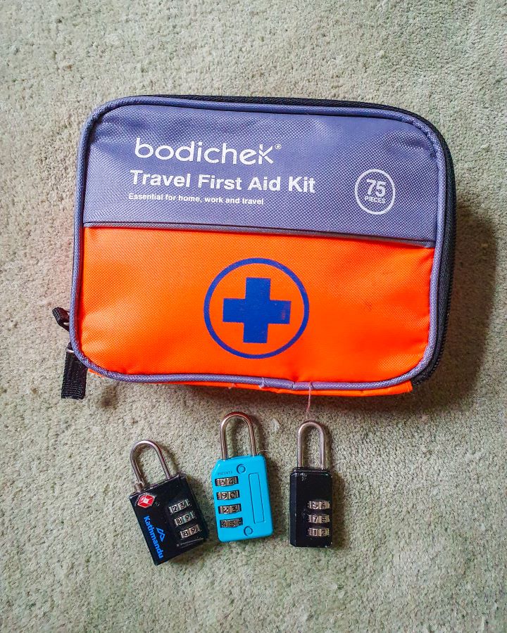 A small orange and grey first aid kit with 75 pieces and 3 little locks underneath it. Two of the locks are black, one is blue and they've got number codes on them to lock them "The Ultimate Travel Packing Guide: 39 Essentials for Every Trip"