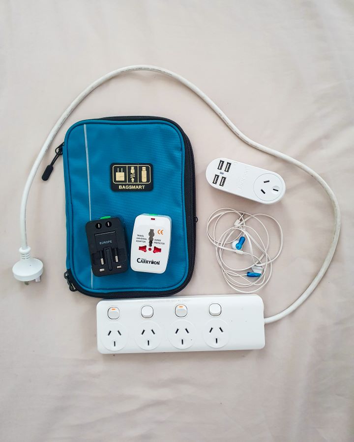 A thin blue cord organiser on the floor with a power board that has 4 outlets, some wired headphones, a white wall charger with one wall outlet and 4 USB slots, and 2 universal adaptors "The Ultimate Travel Packing Guide: 39 Essentials for Every Trip"