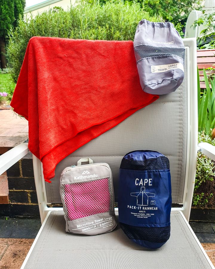 A chair outside with a small red towel folded on it with the packing case next to it. Under them is a small Kathmandu bag with a pink towel folded up inside it and a raincoat folded into a bag beside it "The Ultimate Travel Packing Guide: 39 Essentials for Every Trip"