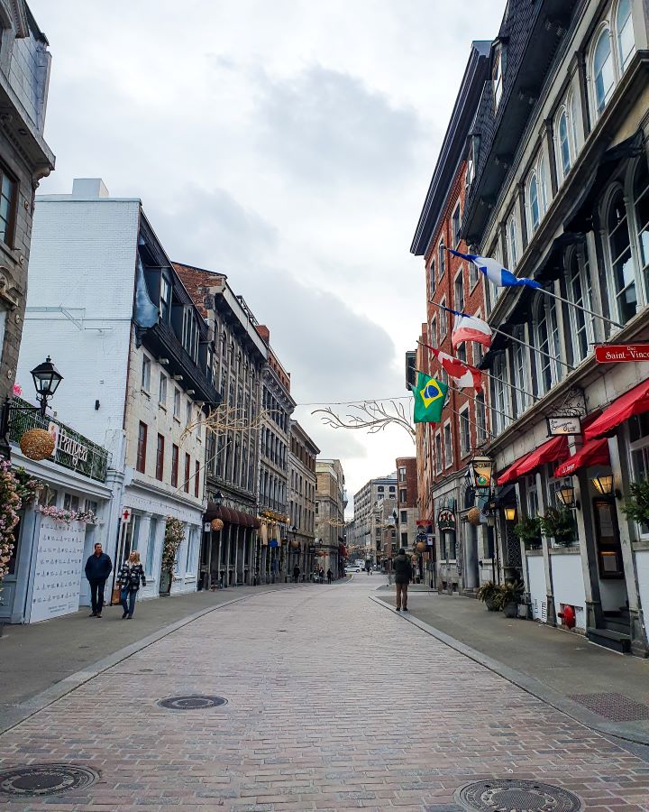 A cute brick street in old town Montréal, Canada. On either side of the street are some lovely buildings with some flags of the world out the front and some old style lamp posts. There are a couple of people on the street but it's mostly pretty empty and there's a decoration of two trees going over the street "Eight Great Free Things To See In Montréal"