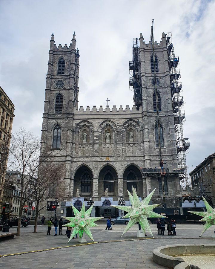 The Notre-Dame Basilica in Montréal, Canada. It's a grey building with two bell towers going up on either side. Out the front are some green star Christmas decorations with some trees and people standing around "Eight Great Free Things To See In Montréal"