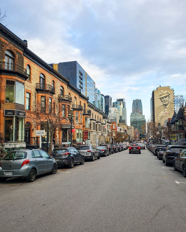 A street in downtown Montréal, Canada with some nice 3 level orange buildings on the left. There are cars parked on both sides of the street with some tall buildings in the distance including a building with some huge art of a man on it "Eight Great Free Things To See In Montréal"