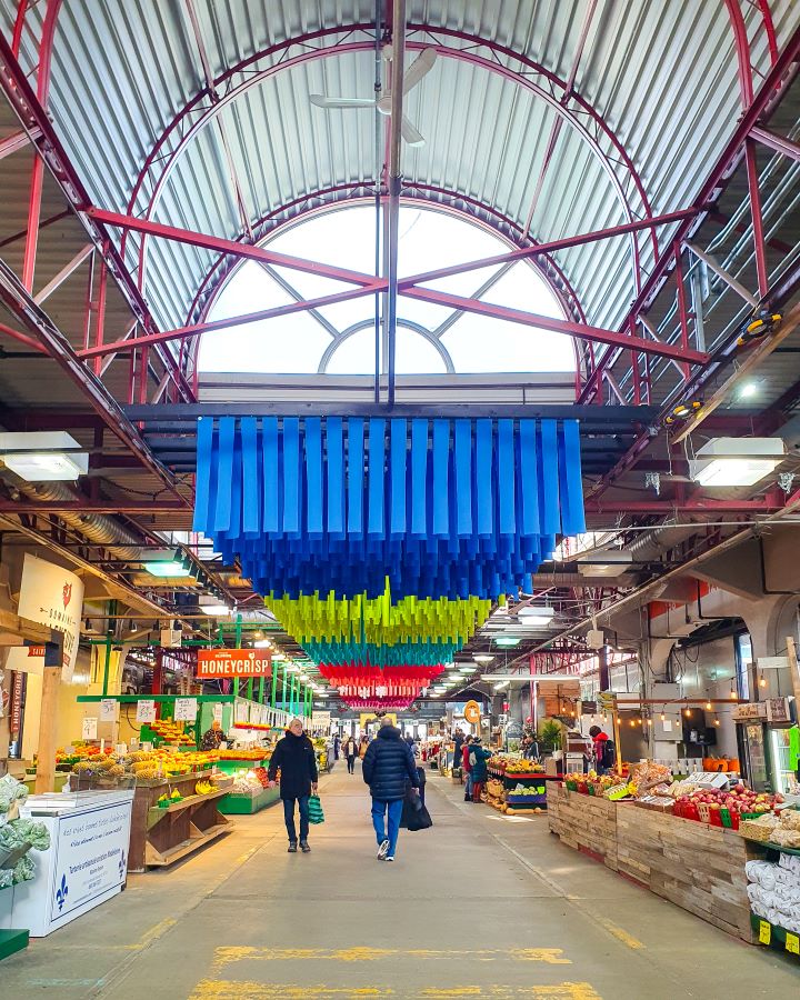 Inside the Jean-Talon Market in Little Italy, Montréal, Canada. There's a big walking space with stalls on either side selling fresh fruit and vegetables. Above the walkway are lots of coloured ribbons and a big arched roof with a glass window "Eight Great Free Things To See In Montréal"