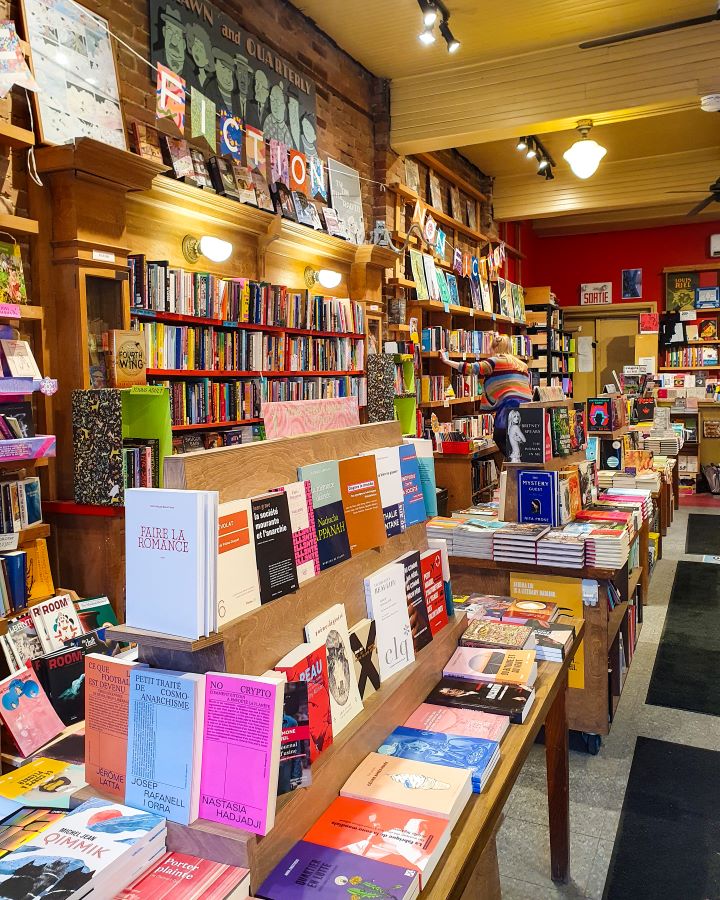 Inside a bookshop in Mile End, Montréal, Canada. There's lots of colourful books everywhere on tables and on shelves along the walls and there's a person shelving in the background "Eight Great Free Things To See In Montréal"