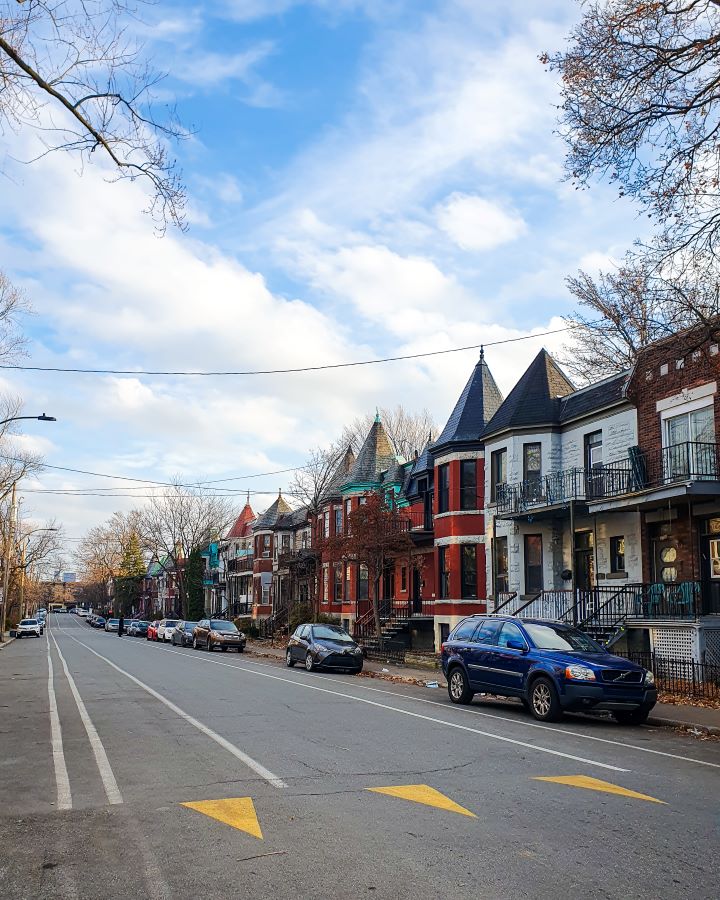 A street in Mile End, Montréal, Canada. On the right side of the street are some very cute houses with some of the red and white houses having pointed roofs. There are also cars and trees on the street and the sky is blue and cloudy "Eight Great Free Things To See In Montréal"