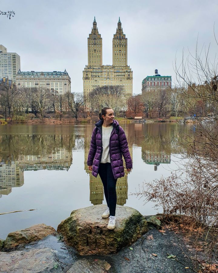 Non-Touristy Activities for Return Visitors to New York City. Krissie standing on a rock with a lake behind her in Central Park, New York City USA. Behind the lake is the famous San Remo building which is a large beige building with two towers on it that end in points. Around the San Remo are more buildings and the buildings reflections are in the lake too