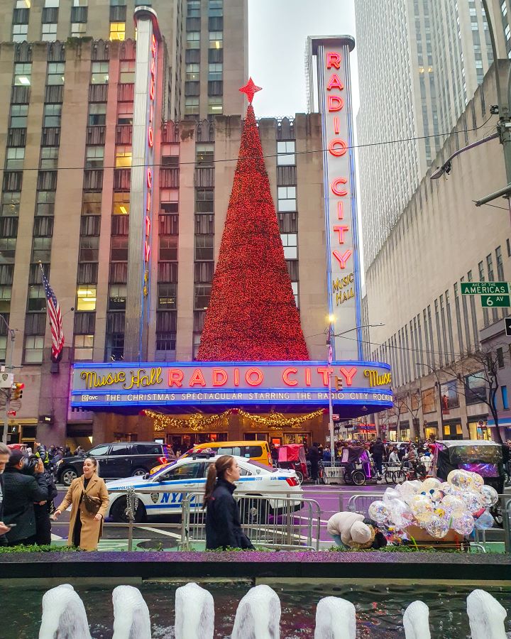 Radio City Music Hall on 6th Avenue in New York City, USA. Radio City has a couple of big signs on the front in yellow and red lights and the big tree on top is red with a big red star on top. In front of it is a road with cars and lots of people around. In the very front of the photo is a fountain with some water spouts "The Ultimate Guide for Musical Theatre Fans In New York"