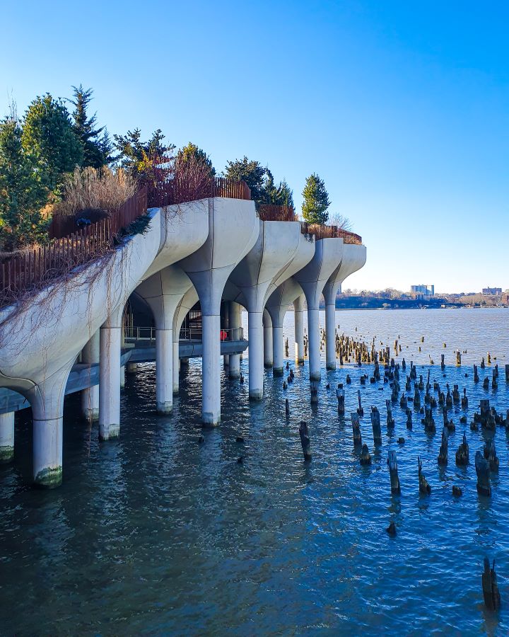 On the left is lots of concrete stands that look a bit like tulips holding Little Island on the Hudson River in New York City, USA. On the right are lots of bits of wooden poles leftover in the water and above it all is a clear blue sky. On Little Island are some trees and bushes and there's a meal railing running along the whole thing "Non-Touristy Activities for Return Visitors to New York City"