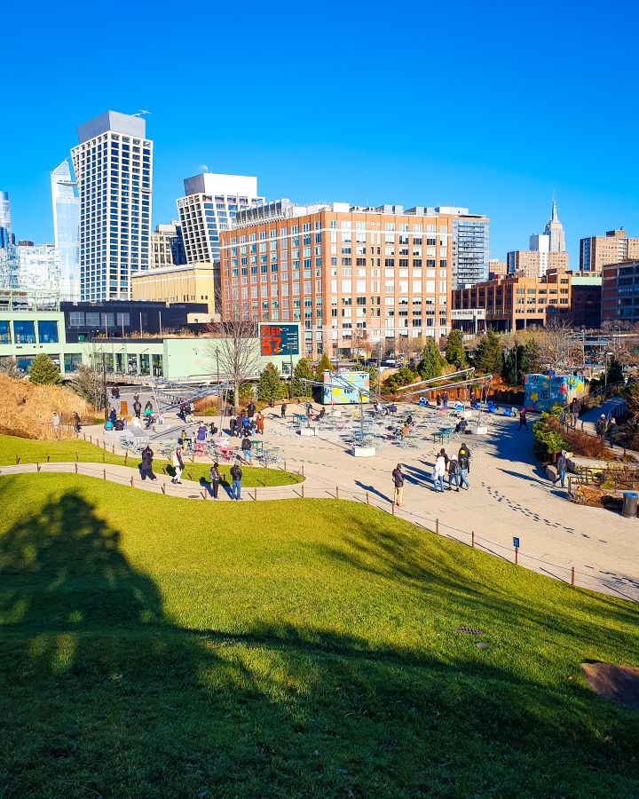 On Little Island in New York City, USA. There's a large space of short green grass with a railing around it and a path behind it. There's people walking on the path as well as seating areas and more plants. Behind the island is a clear view of a part of Manhattan with lots of buildings and the Empire State Building in the distance "Non-Touristy Activities for Return Visitors to New York City"