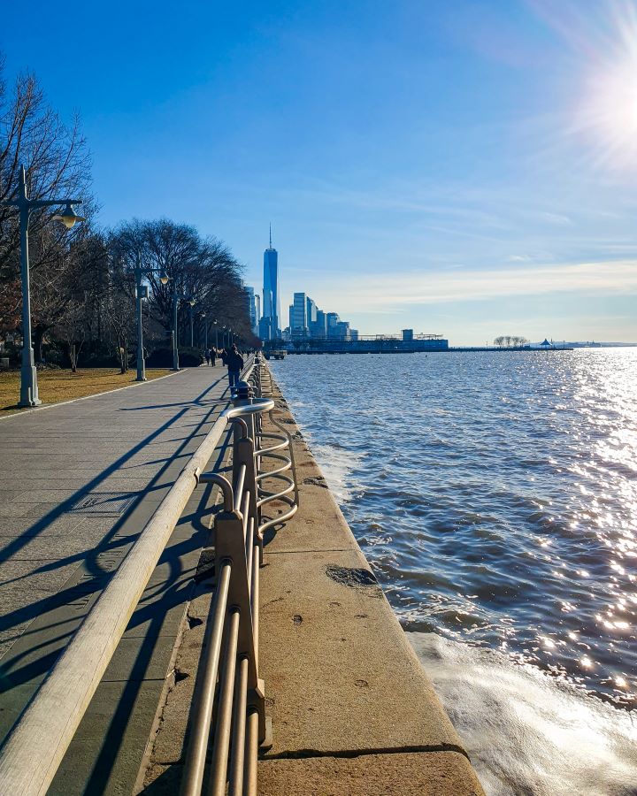 Looking down along the Hudson River in New York City, USA. On the left is a path with a metal railing alongside it with some light poles and and trees along the side of the path. On the right is the blue water with the blue sky above it all and in the distance are some tall high rise buildings at the bottom of Manhattan "Non-Touristy Activities for Return Visitors to New York City"