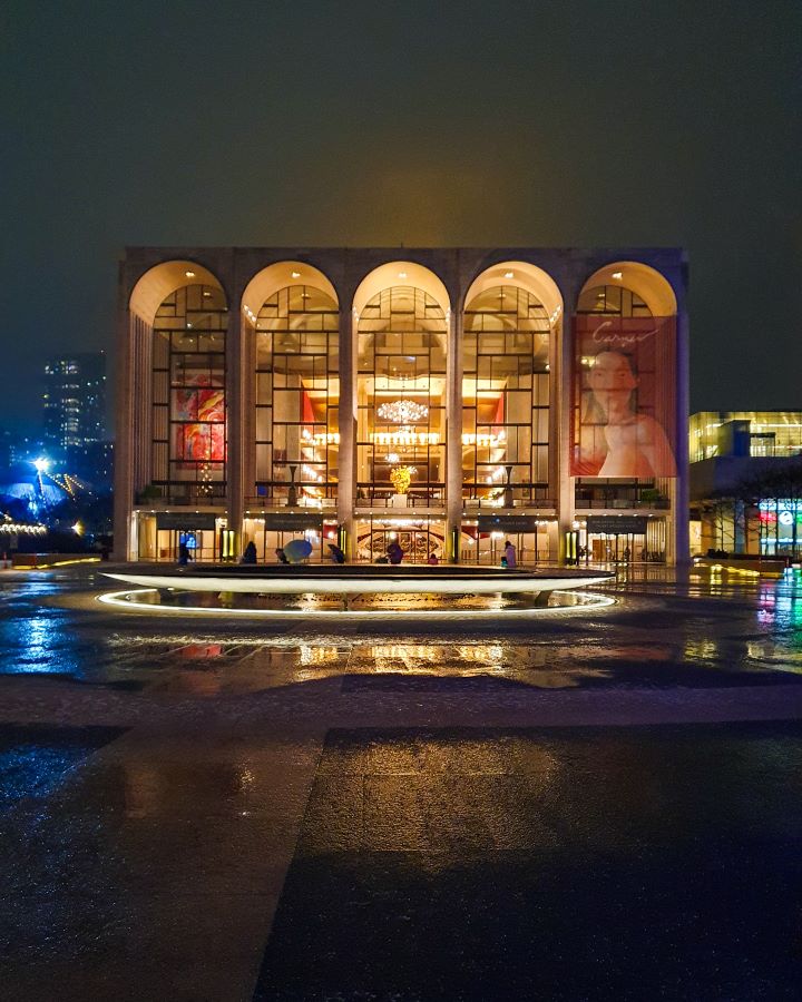 Lincoln Centre in New York City, USA. The photo is taken at night and Lincoln Centre is lit up with 5 huge arches at the front with glass windows showing the warm light inside. Out the front is a fountain also lit up and the rest of the pavement is dark and wet "The Ultimate Guide for Musical Theatre Fans In New York"