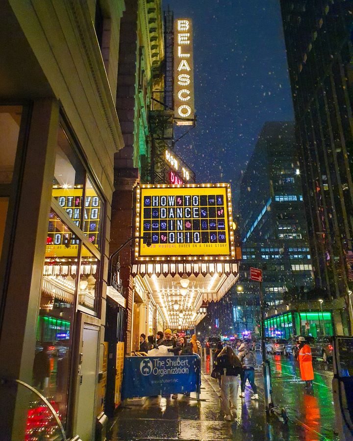 The Belasco Theatre in New York City, USA. There's a big sign lit up at the top that says "Belasco". Underneath there's a marquee that says "How to Dance in Ohio" in a yellow square. Under that are people lining up, and behind them is a road with cars and more buildings behind it. The photo is taken at night and you can see it's lightly snowing! "The Ultimate Guide for Musical Theatre Fans In New York"