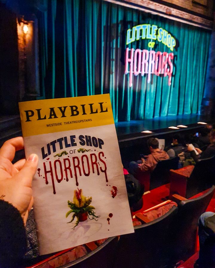 A hand holding a little book that says "Playbill" up the top in a yellow box and "Little Shop of Horrors" on the bottom in New York City, USA. Behind the Playbill are red seats and a stage with a big green curtain that says "Little Shop of Horrors" on it "The Ultimate Guide for Musical Theatre Fans In New York"