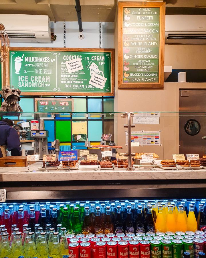 Inside Schmackery's in New York City, USA. In the bottom of the photo are heaps of drinks in bottles and above that is the counter with a glass barrier. Behind it you can see lots of cookies with signs, and behind that are some signs with the cookie flavours "The Ultimate Guide for Musical Theatre Fans In New York"