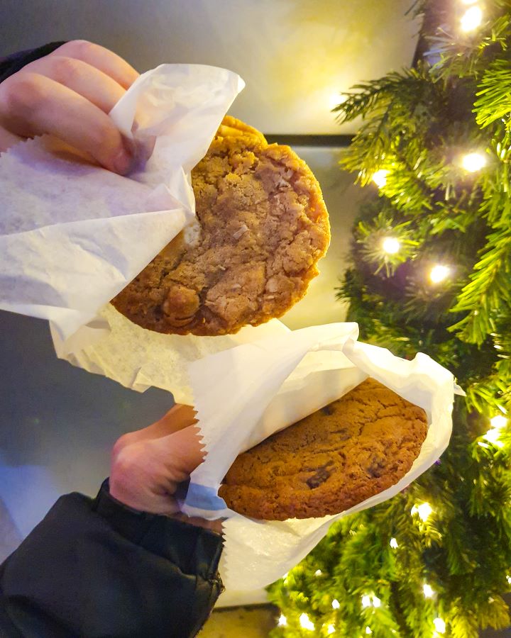 Two hands holding a cookie each in a white bit of paper in New York City, USA. On the right behind the hands is a bit of greenery with some Christmas lights in it "The Ultimate Guide for Musical Theatre Fans In New York"