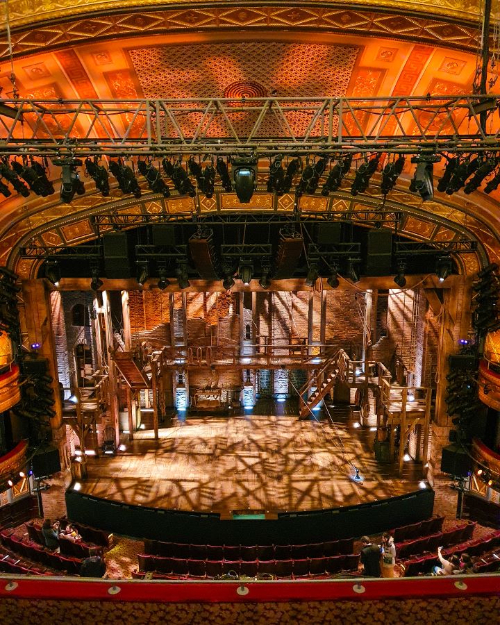 Inside a theatre in New York City, USA. The stage has the set for Hamilton on it which has a wooden banister around the wooden stage and there's lights above the stage. The photo is taken from the second level so it's looking down at the stage from above "The Ultimate Guide for Musical Theatre Fans In New York"