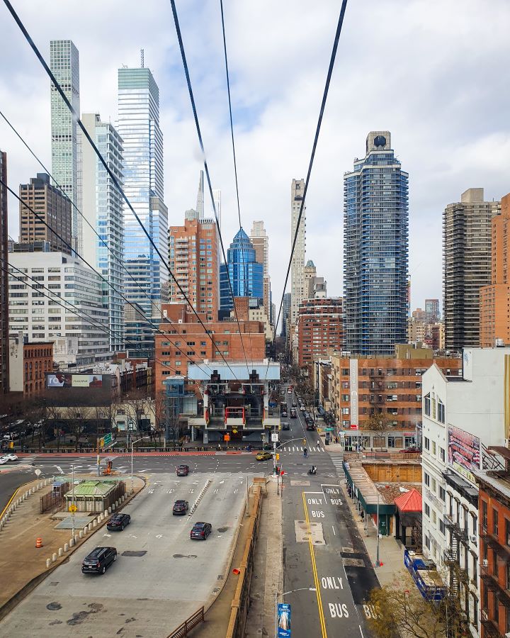 A view from the tramway that goes from Manhattan over to Roosevelt Island in New York City, USA. There's 8 wires coming from the top of the photo leading back to the building the tramway comes from. Around that building are the high rises of Manhattan, roads, cars and people walking around "Non-Touristy Activities for Return Visitors to New York City"