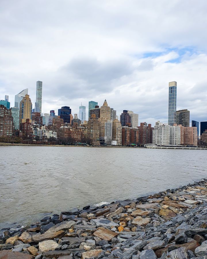 A view back to Manhattan from Roosevelt Island New York City, USA. In the foreground are rocks on the bank of the island, with the brown river behind it. Beyond the river is Manhattan which has lots of high rises and the sky is really cloudy (it ended up snowing this day) "Non-Touristy Activities for Return Visitors to New York City"