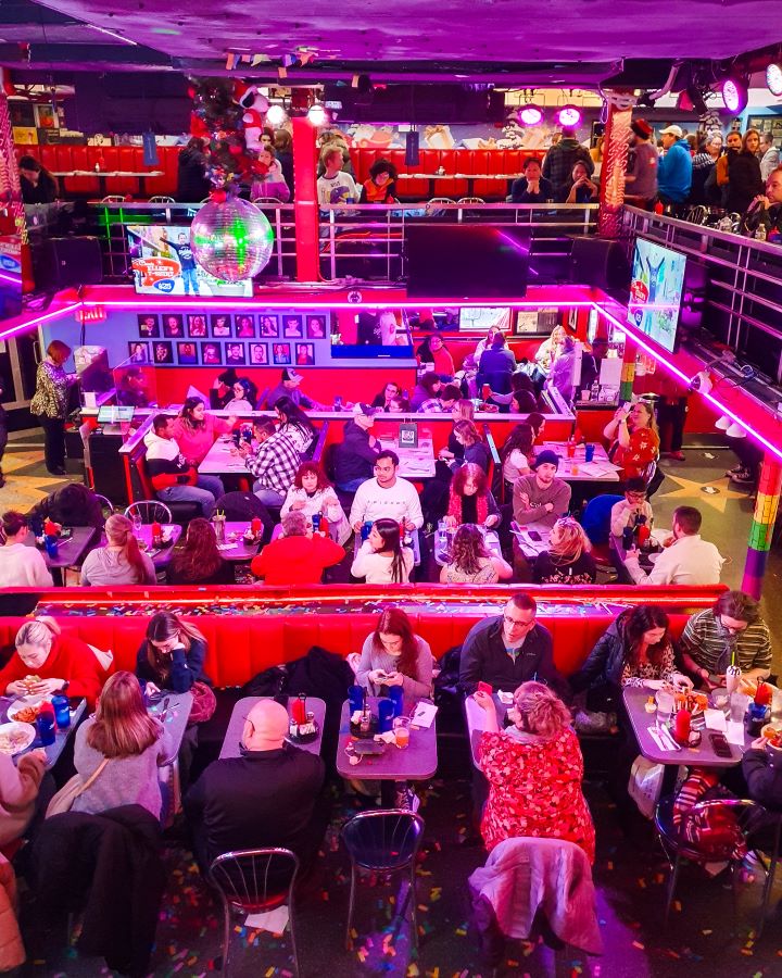 Inside Ellen's Stardust Diner in New York City, USA. The diner is 1950s themed and the photo is taken up on the second level, looking down on the bottom level. There are lots of tables and chairs squished together with lots of people sitting at them. They're booth style and there's lots of red material all around. The top level has more seating and people and there are TVs on the balconies and a disco ball hanging from the roof "The Ultimate Guide for Musical Theatre Fans In New York"