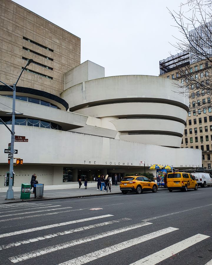 The Guggenheim Museum in New York City, USA. In the foreground is the road with a couple of yellow taxis on it. Behind them is the museum which is a big cream coloured building that has a bowl shape on top with black stripes on it "Non-Touristy Activities for Return Visitors to New York City"