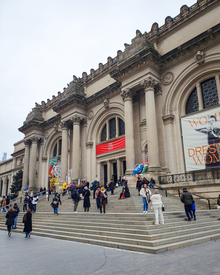 The Met museum in New York City, USA. It's a big beige building with 6 columns out the front and a fancy roof. There's also three big arched windows on the front and stone steps leading up to the doors with people all over the stairs "Non-Touristy Activities for Return Visitors to New York City"